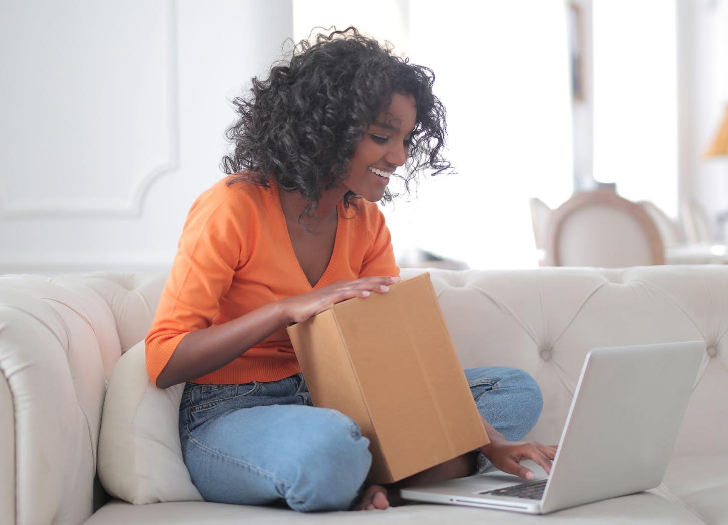 Black woman smiling with a cardboard box in her lap as she types on a laptop